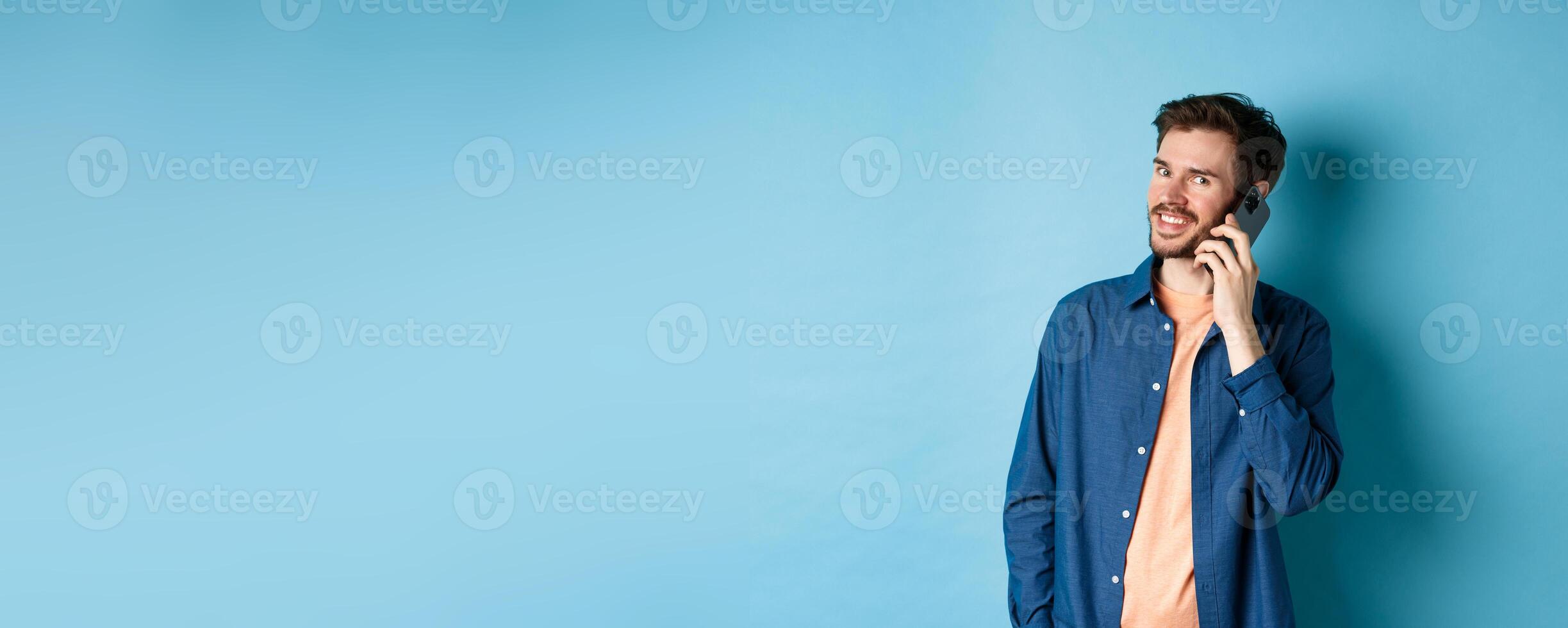 Modern smiling guy talking on mobile phone, looking happy at camera, standing on blue background photo