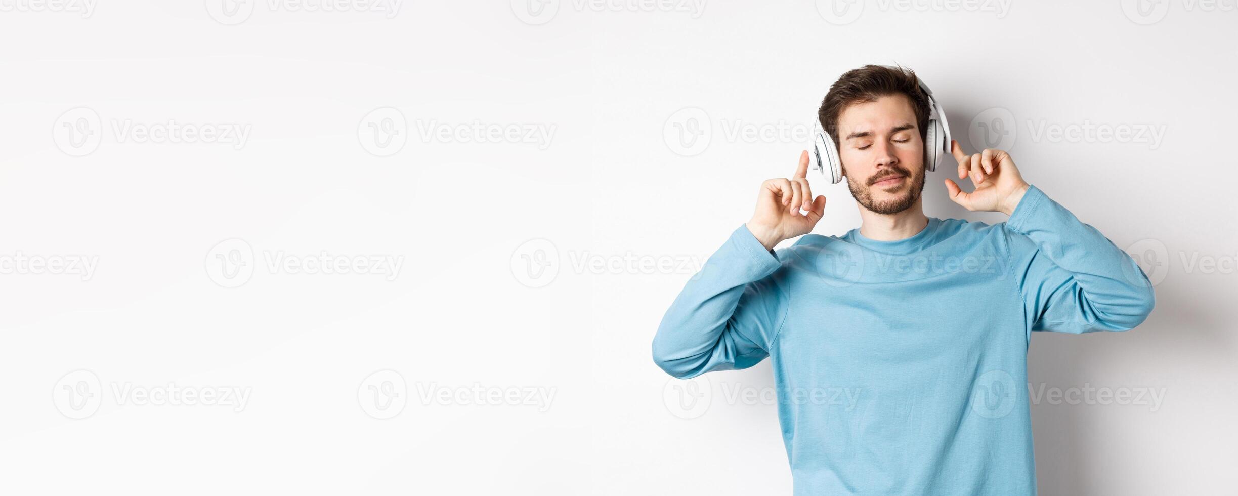 Relaxed young man enjoying favorite song, listening music in headphones with closed eyes and calm face, standing over white background photo
