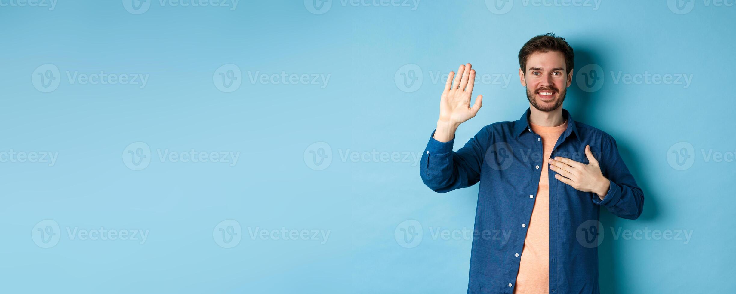 Happy young man introduce himself, telling truth with hand on heart and friendly smile, standing on blue background photo