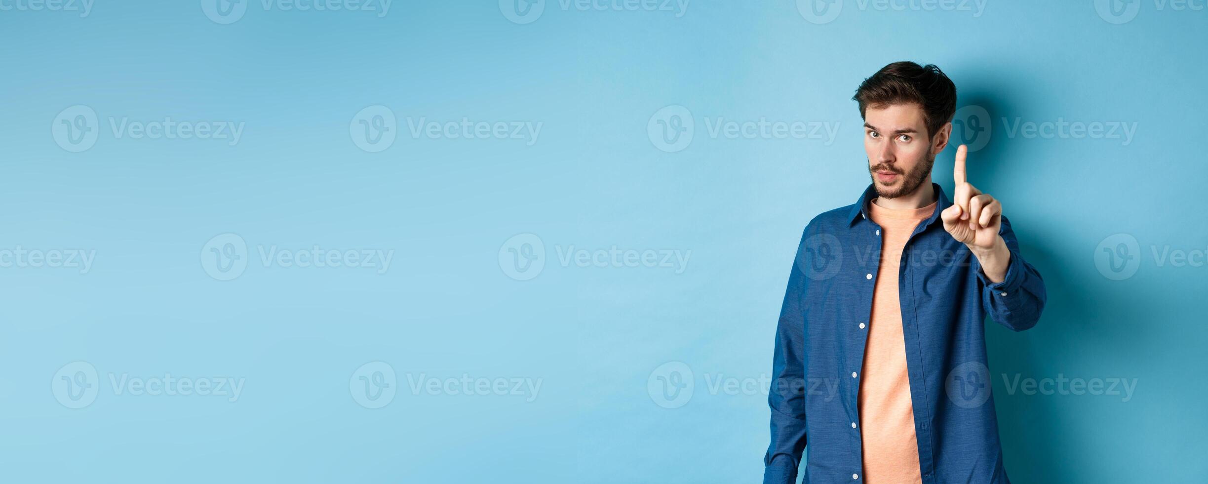 Image of young man give warning, teaching a lesson, raising one finger to scold, looking at camera, patronizing someone, standing on blue background photo