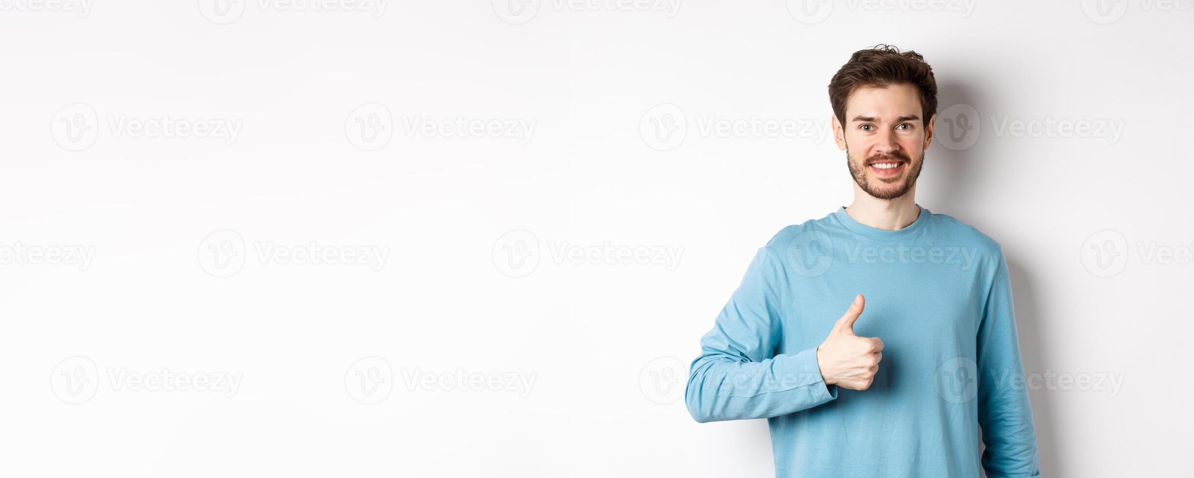 Smiling male model in casual shirt showing thumb up, approve and recommend product, praise excellent choice, standing on white background photo