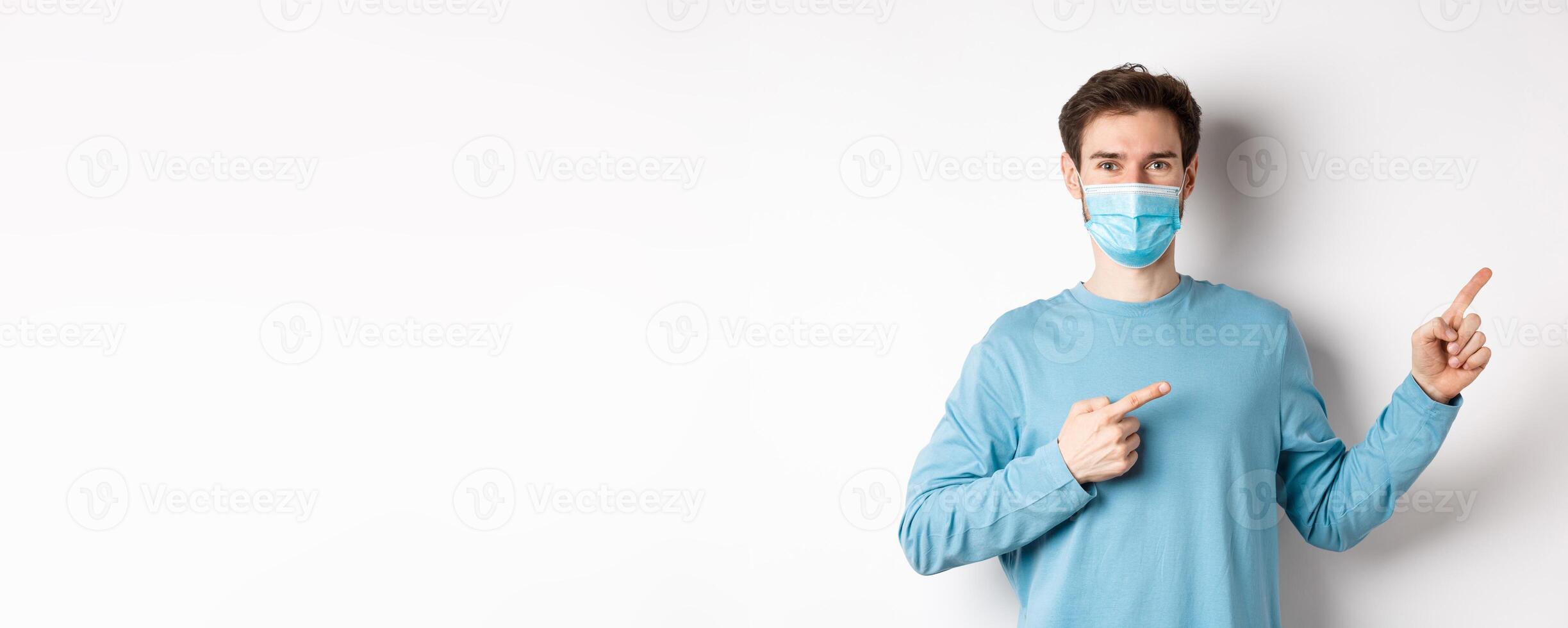 Coronavirus, health and quarantine concept. Handsome caucasian man in face mask pointing fingers right, showing advertisement, standing over white background photo