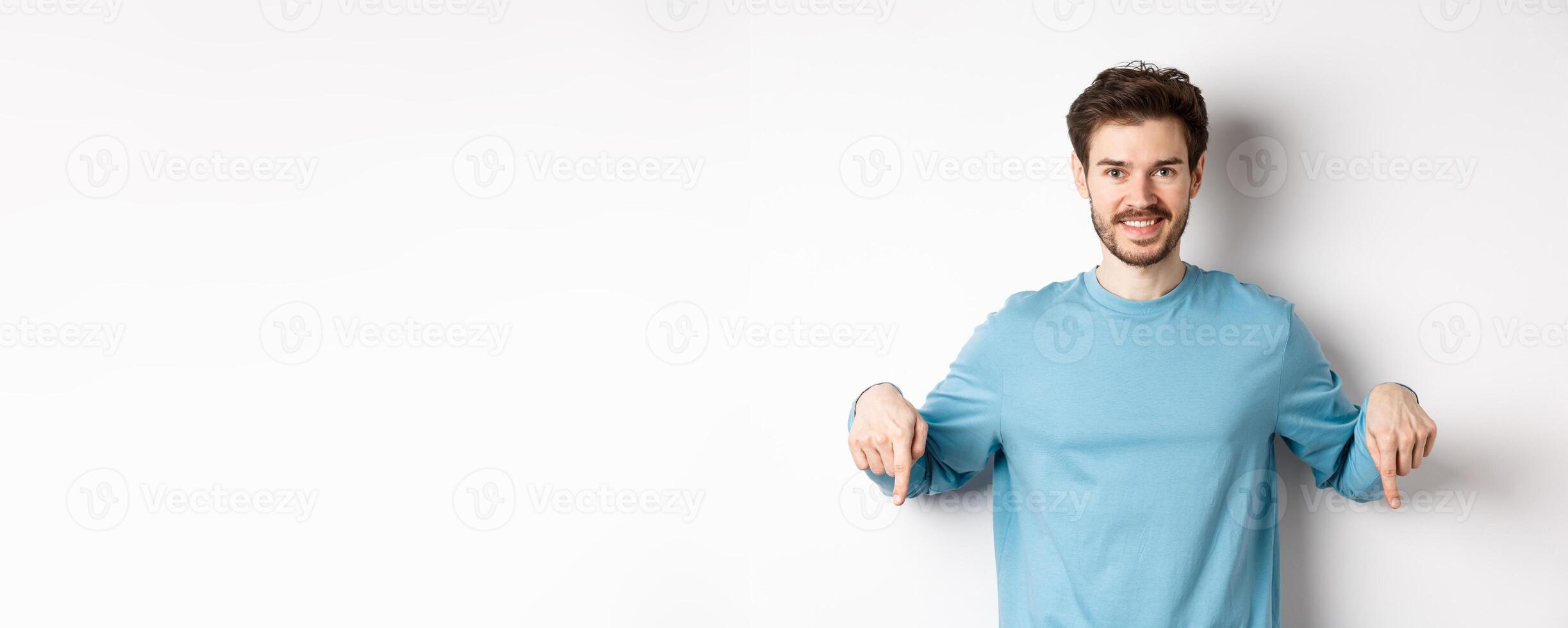 Smiling confident man pointing fingers down, showing promo banner or logo on white background, standing in casual blue sweatshirt photo