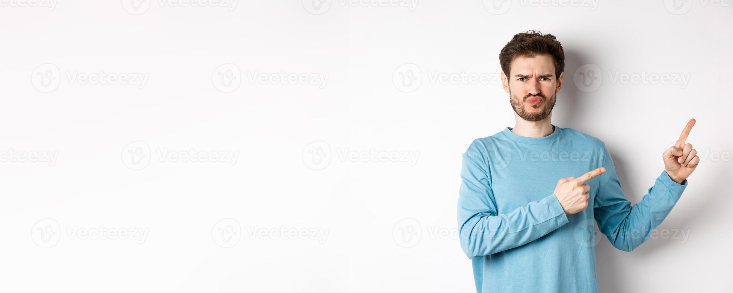 Disappointed and upset young man showing bad offer, pointing fingers right and frowning disgusted, standing with skeptical face on white background photo