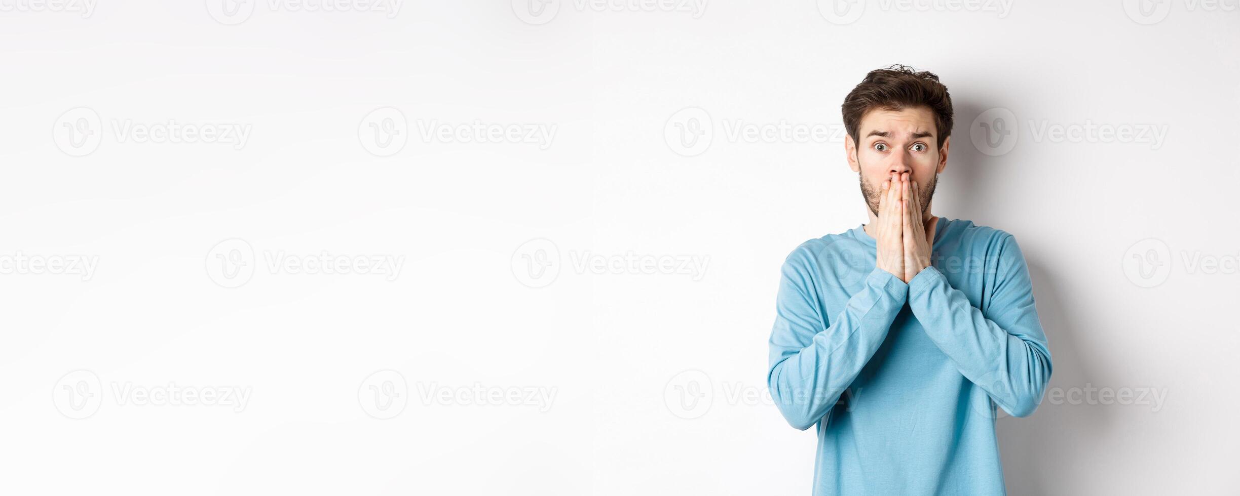 Shocked and worried young man gasping and covering mouth, look with panic at camera, standing anxious against white background photo