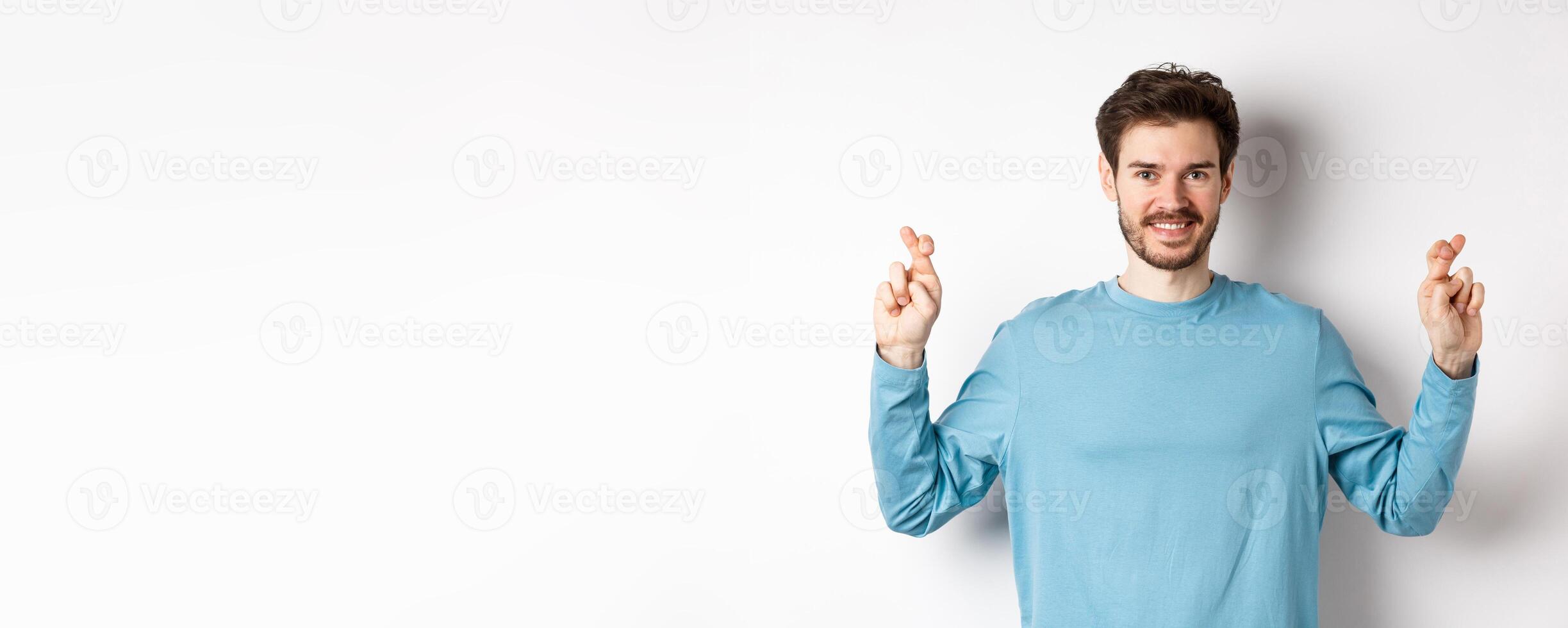 Optimistic smiling man cross fingers for good luck, making wish and waiting for results, standing over white background photo