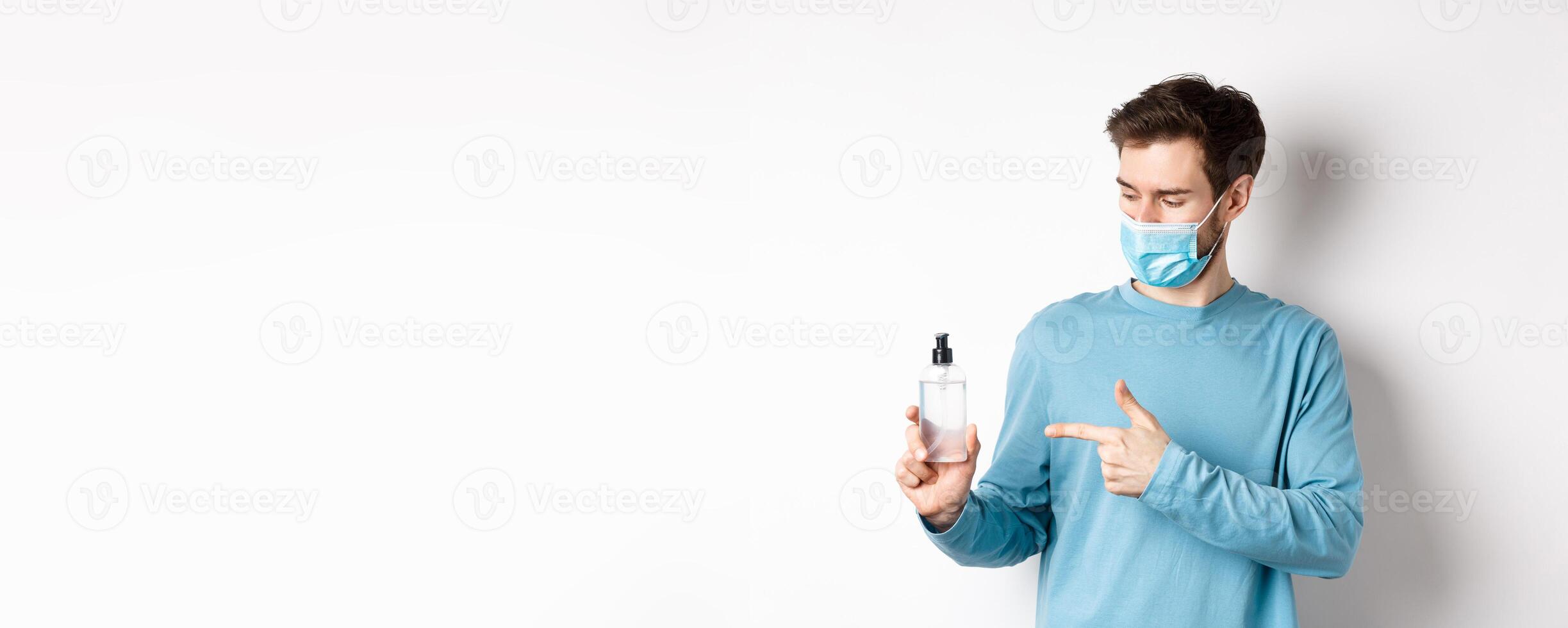 Covid-19, health and quarantine concept. Young man pointing and looking at hand sanitizer bottle, showing good antiseptic, white background photo