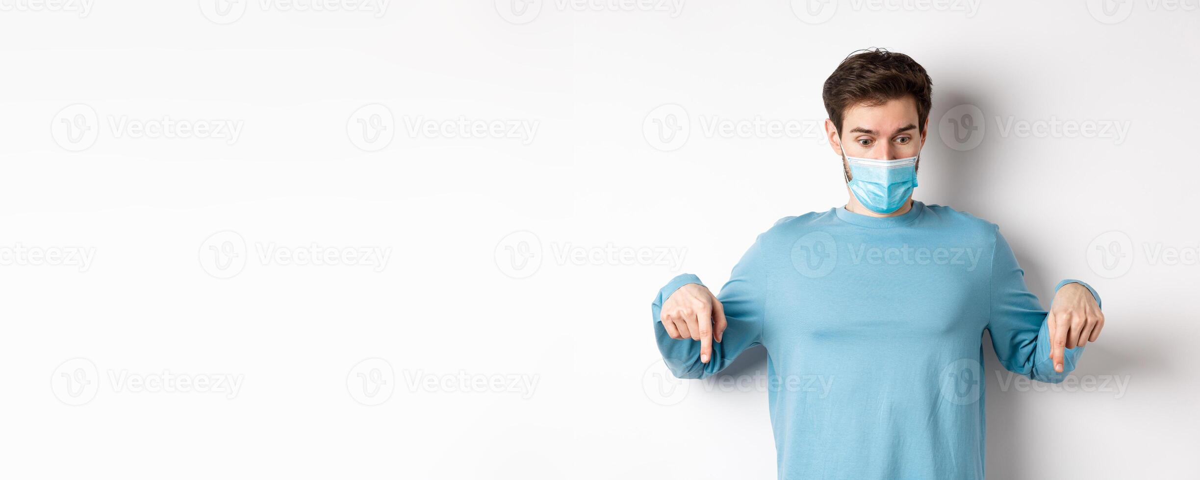 Covid-19, health and quarantine concept. Curious man checking out advertisement, pointing and looking down, wearing medical mask with casual blue sweatshirt, white background photo