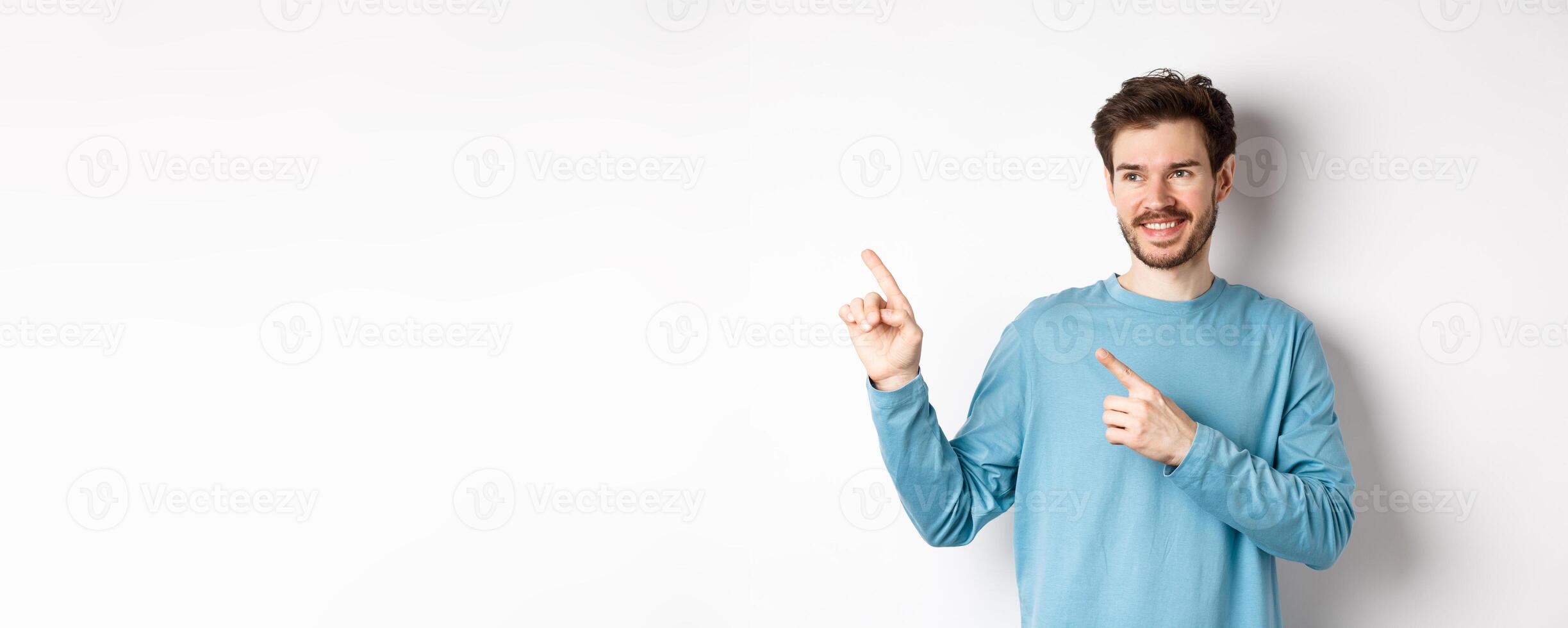 Portrait of young modern man in blue sweatshirt pointing fingers at upper left corner and looking away, smiling pleased as showing advertisement on white background photo