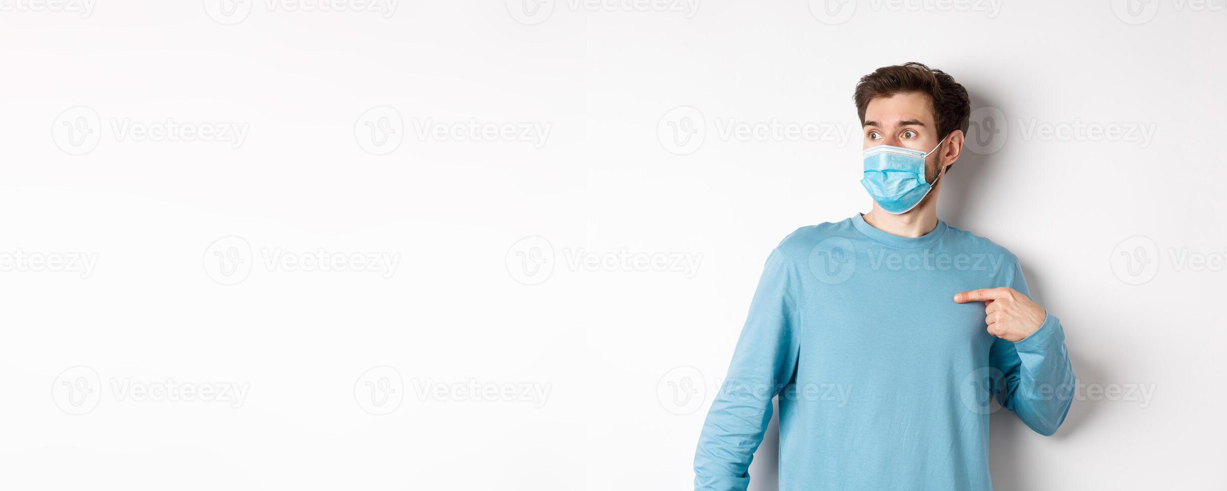 Covid-19, health and quarantine concept. Confused man in medical mask pointing at himself, looking left with ambushed face, standing over white background photo