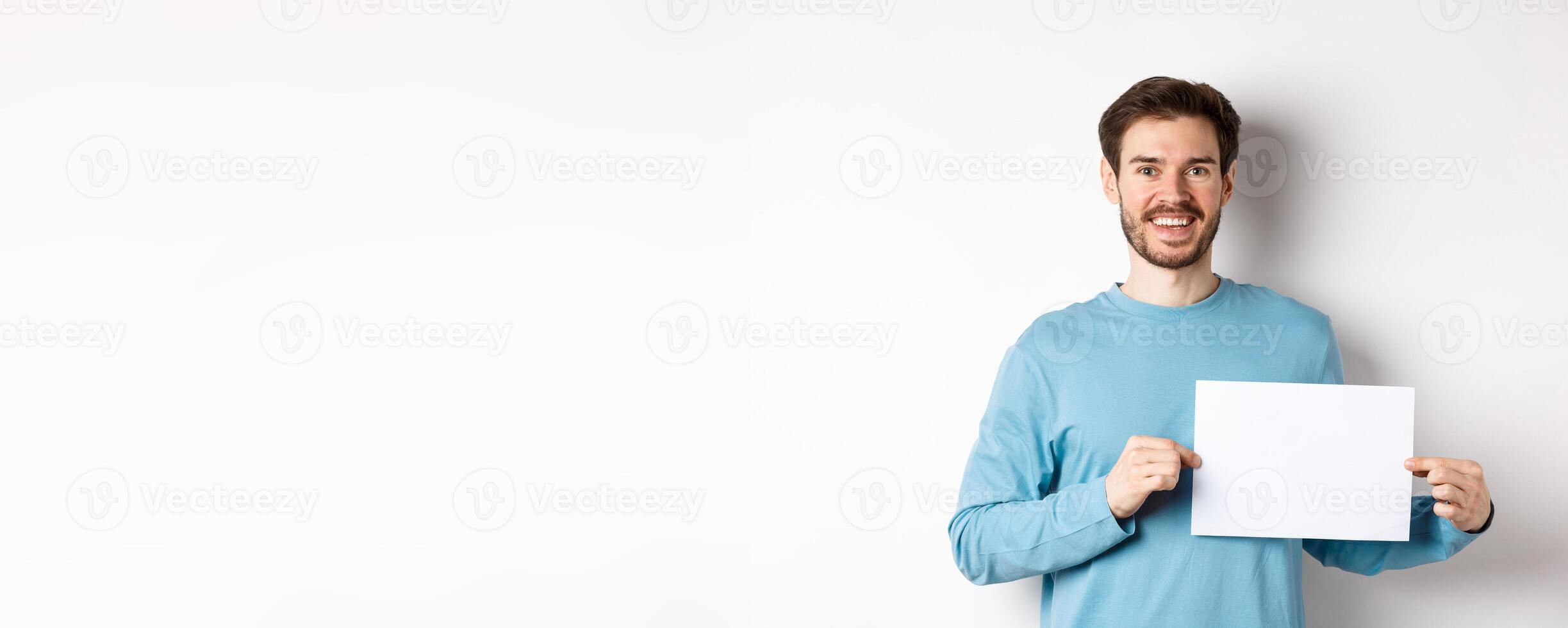 Young man with happy face showing blank piece of paper for your logo or sign, smiling at camera, standing over white background photo