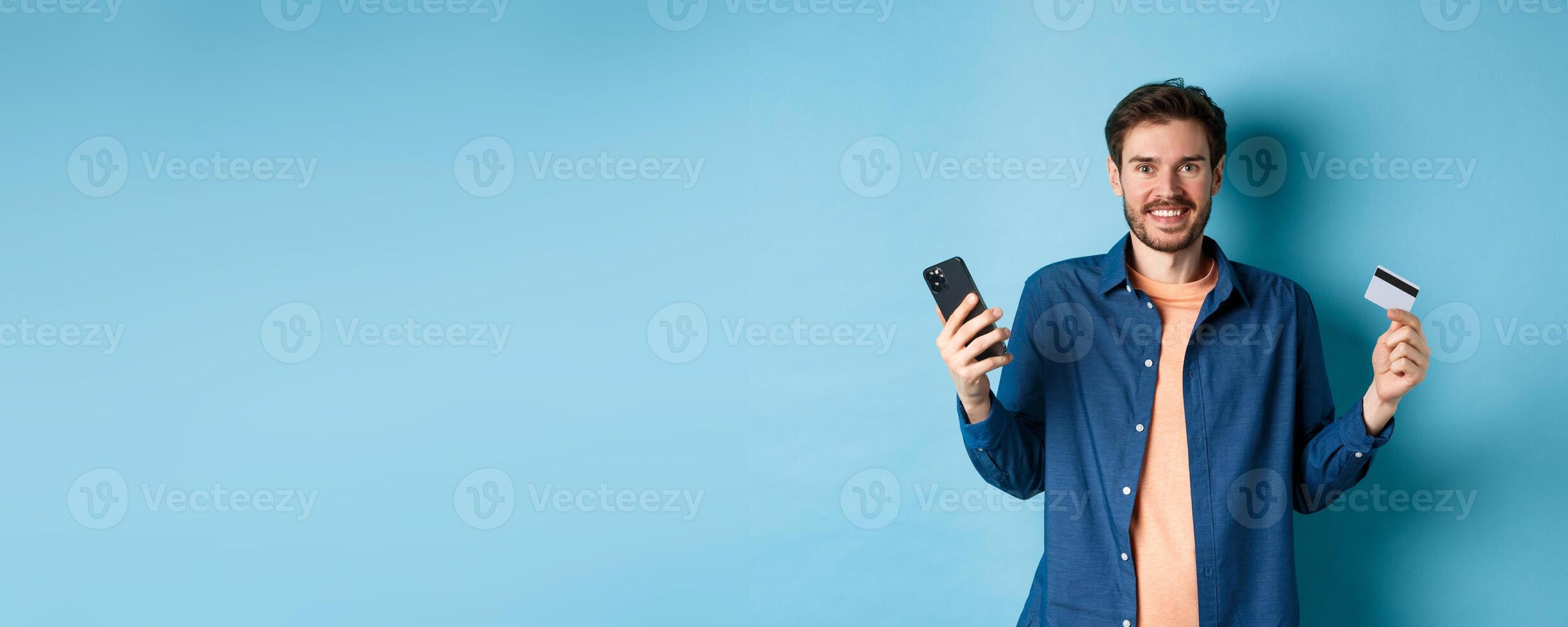 E-commerce concept. Excited young man holding smartphone and plastic credit card, shopping online, standing on blue background photo