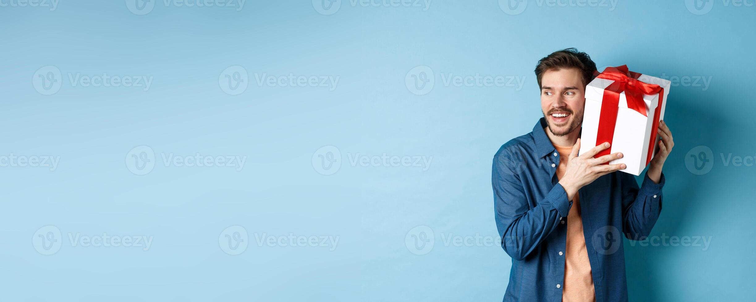Valentines day. Smiling man guessing what inside gift box, got present from lover girlfriend, standing against blue background photo