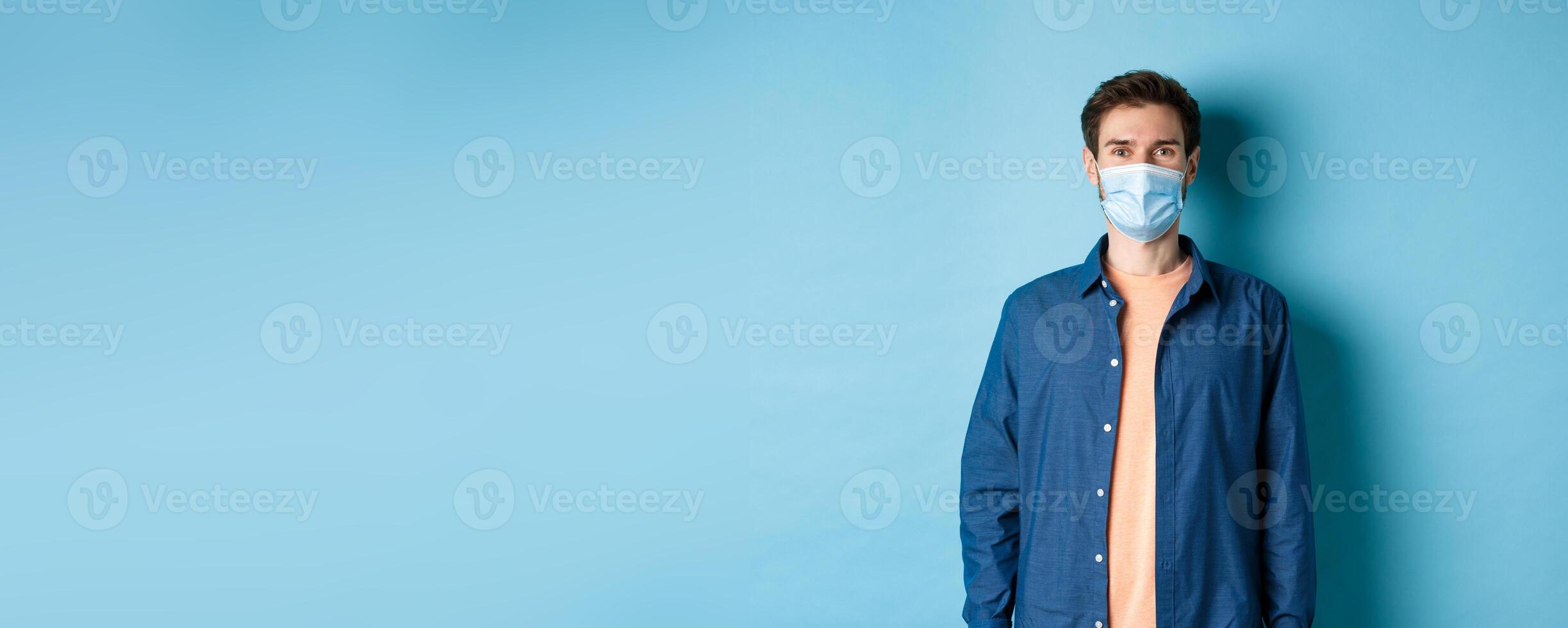 Covid-19 and healthcare concept. Smiling young man in medical mask looking healthy and happy, standing on blue background photo