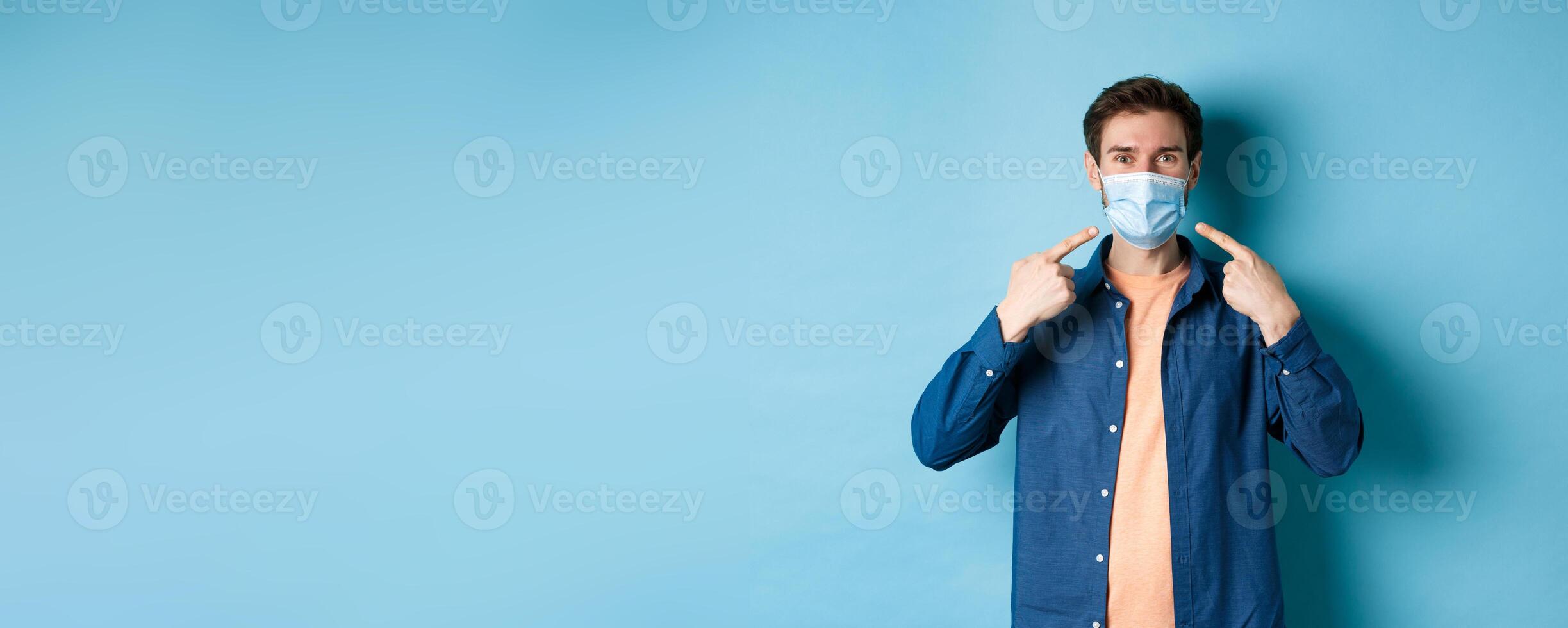Coronavirus and pandemic concept. Portrait of happy and healthy man pointing fingers at his medical mask, smiling at camera, standing on blue background photo