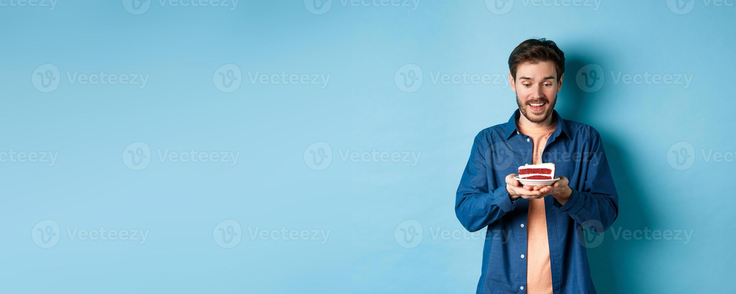 Celebration and holiday concept. Happy young man looking at birthday cake and making wish, standing on blue background photo