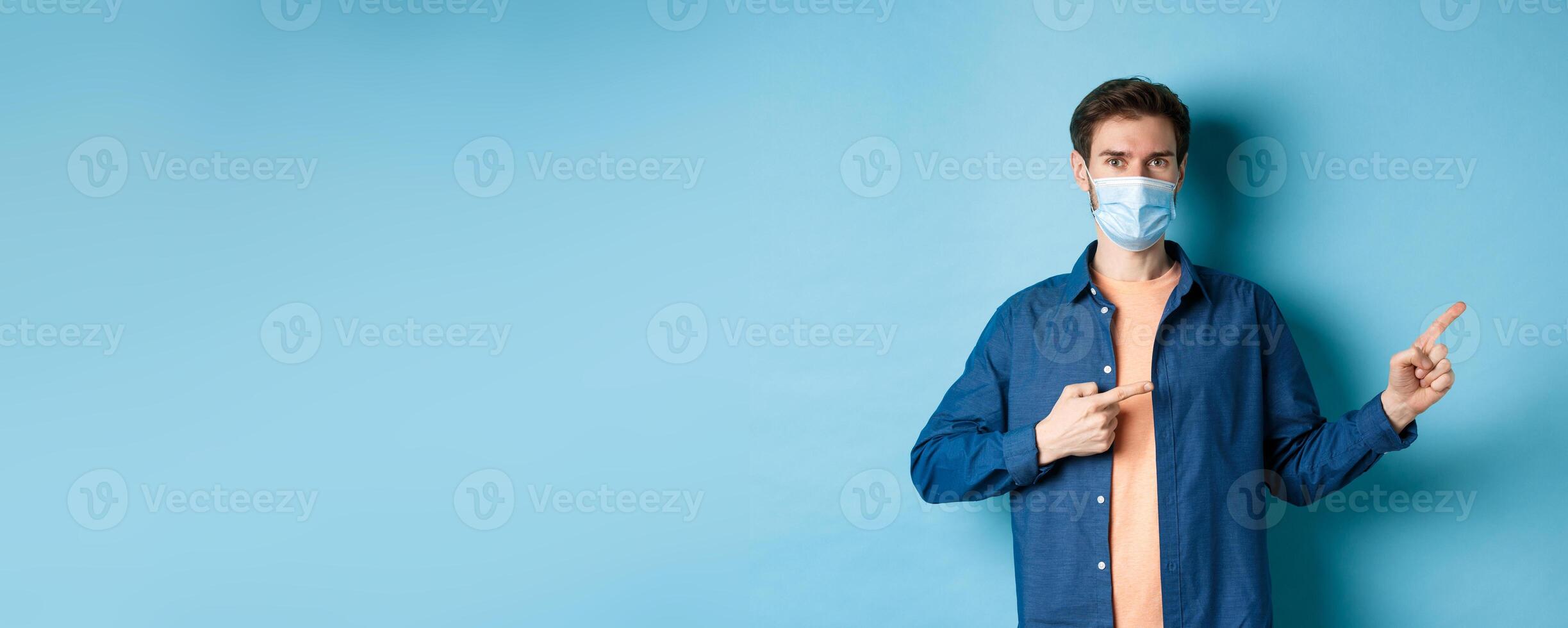 Covid and healthcare concept. Handsome modern guy in medical mask pointing at empty space aside, standing on blue background photo