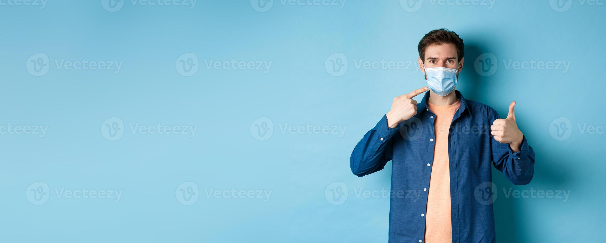 Coronavirus and pandemic concept. Young healthy man pointing at medical mask and showing thumbs up, using preventive measures from catching covid-19, blue background photo