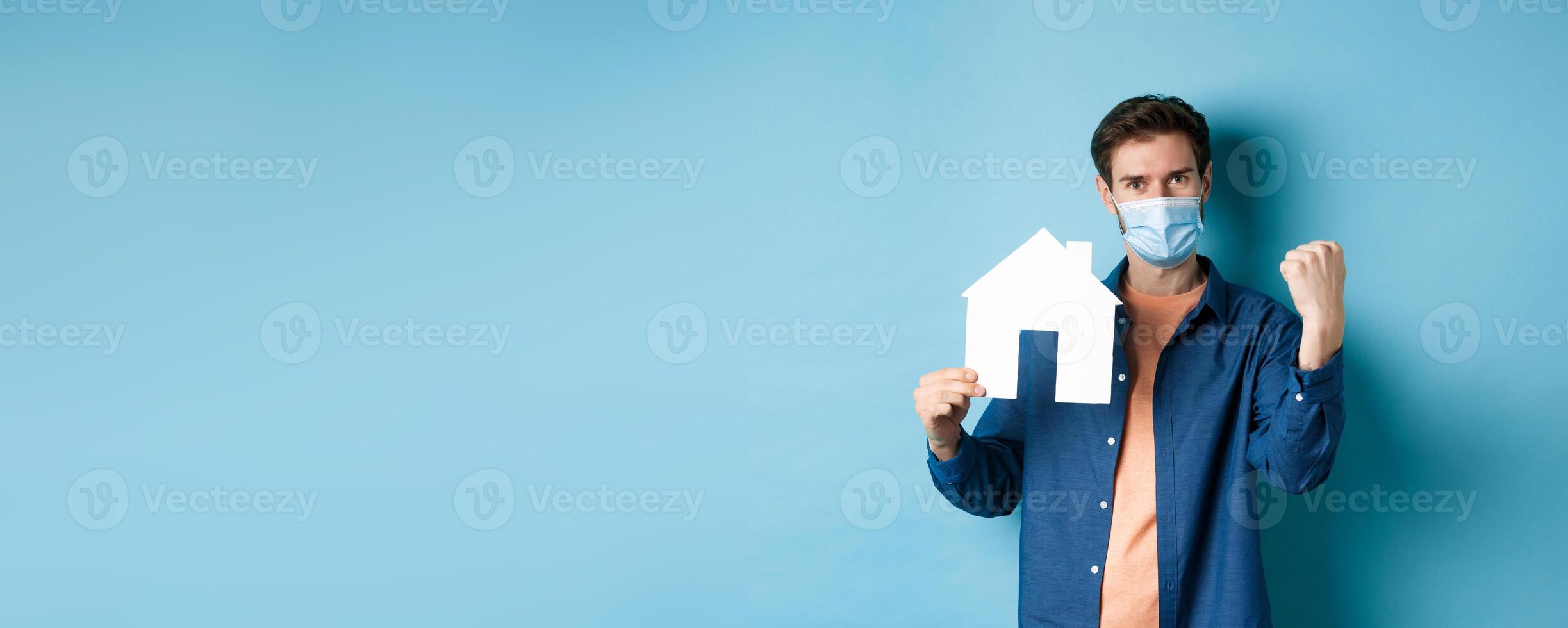 Real estate and covid concept. Cheerful guy in medical mask showing paper house cutout and fist pump, buying property and celebrating, standing on blue background photo