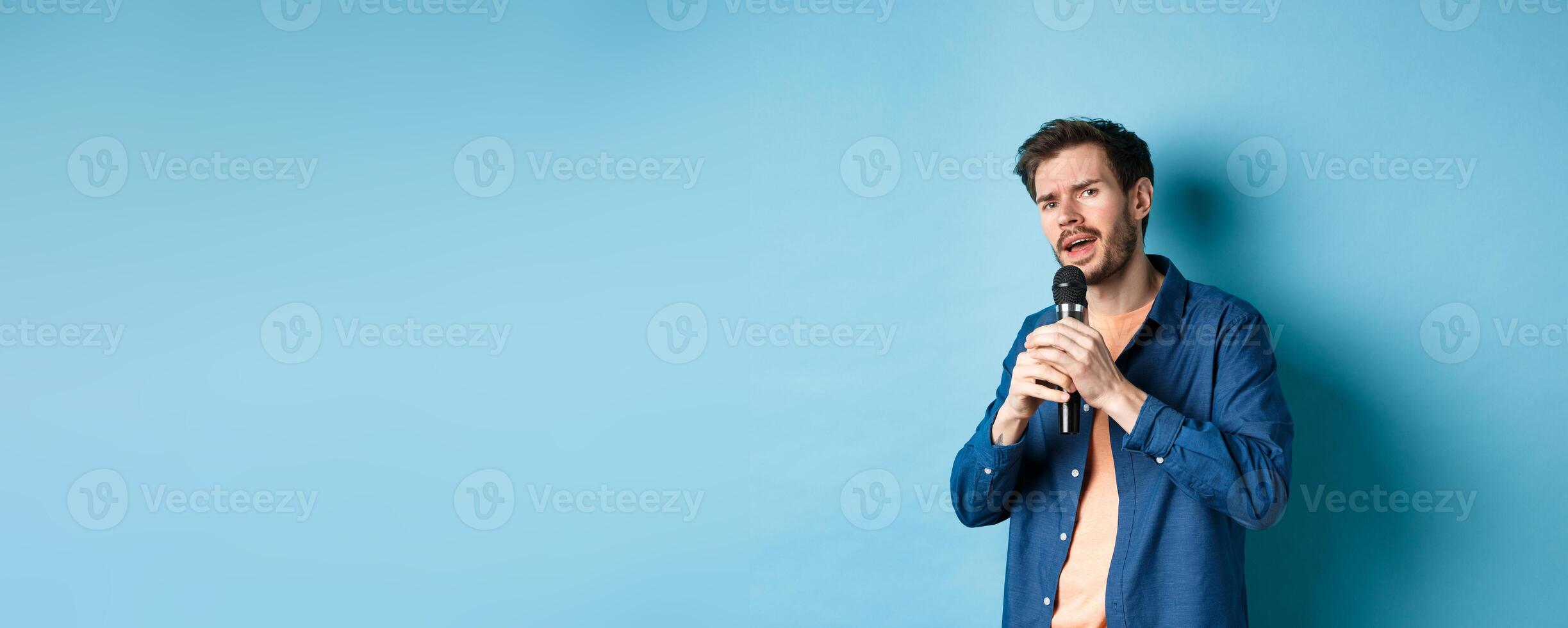 Young man singing song in microphone, playing karaoke, standing on blue background photo