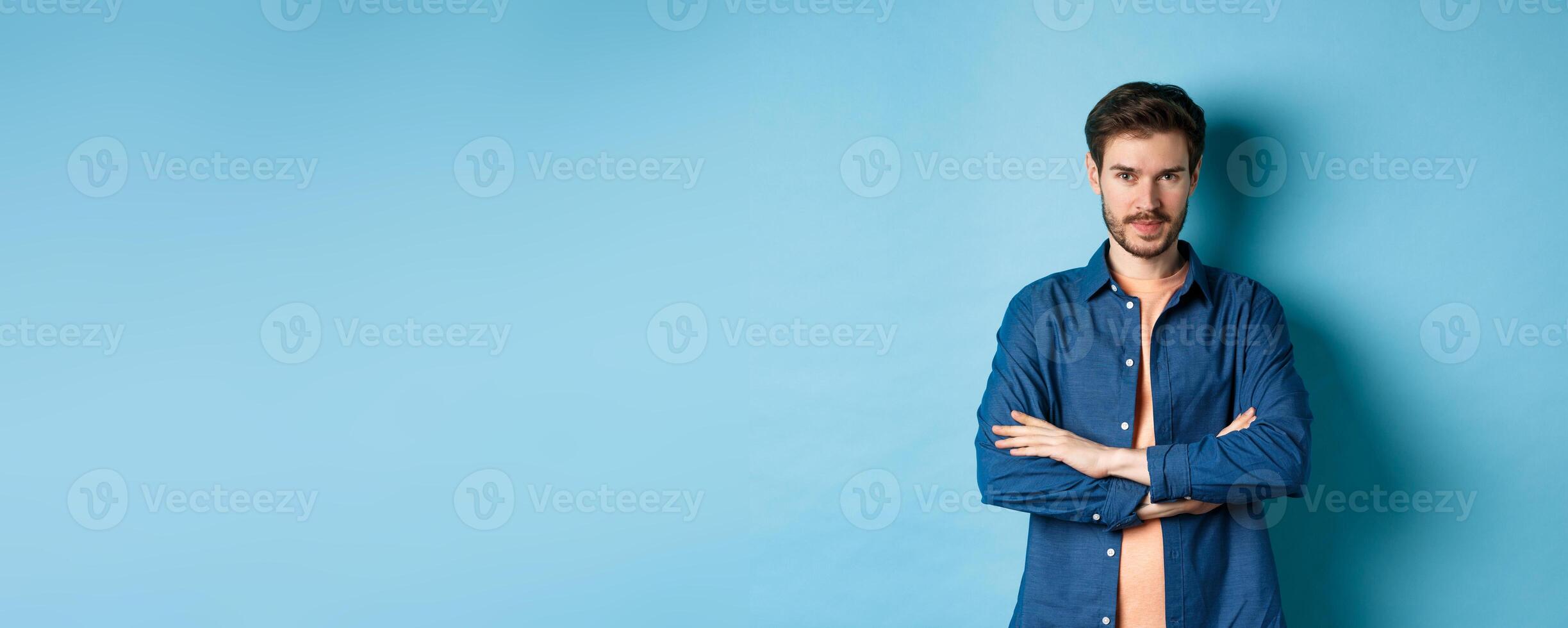 Confident young man feeling like professional, cross arms on chest and smiling sassy, standing on blue background photo