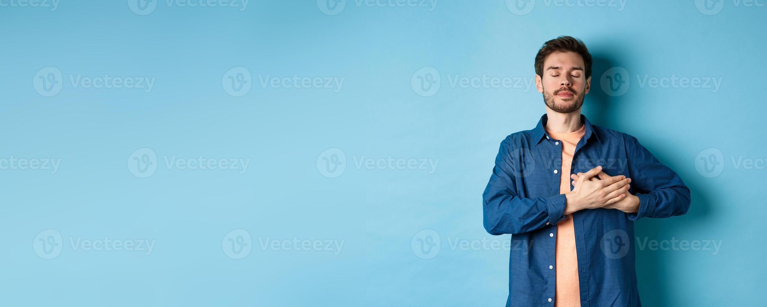 Romantic and nostalgic guy holding hands on heart with eyes closed, keep warm memories, standing on blue background photo