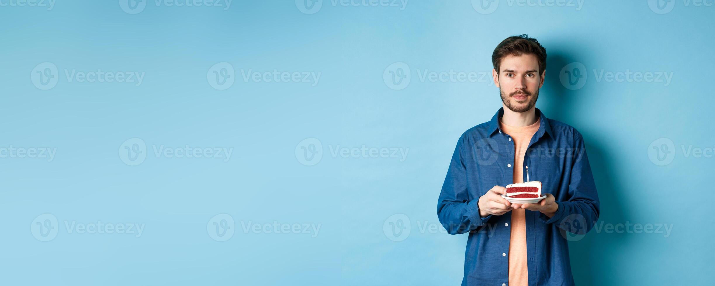 Image of smiling young man celebrating birthday, holding b-day cake with lit candle and looking at camera, standing over blue background photo