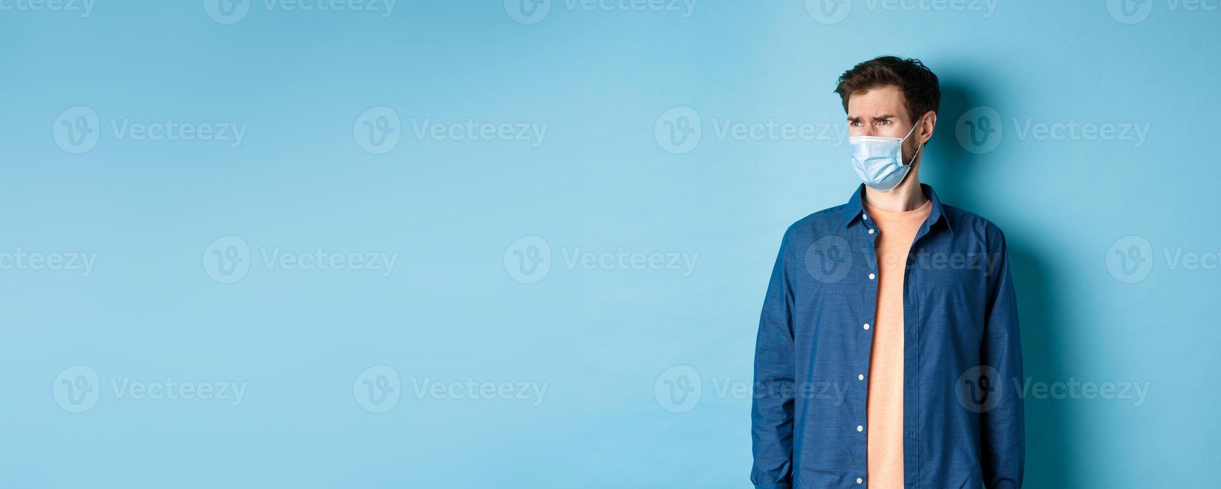 Covid-19 and healthcare concept. Dissatisfied frowning guy in medical mask looking at bad promo, stare aside at empty space with disappointed face, blue background photo
