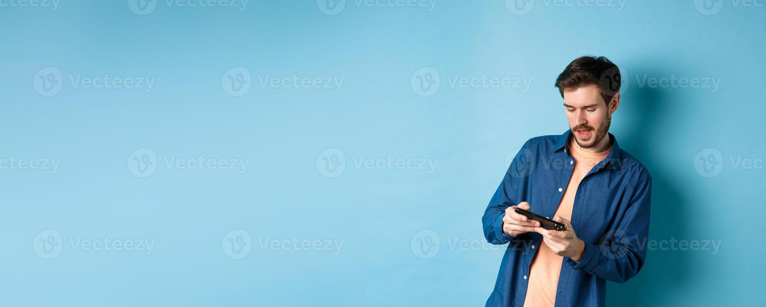 Young guy playing game on mobile phone, tilt body and holding smartphone horizontally, standing on blue background photo
