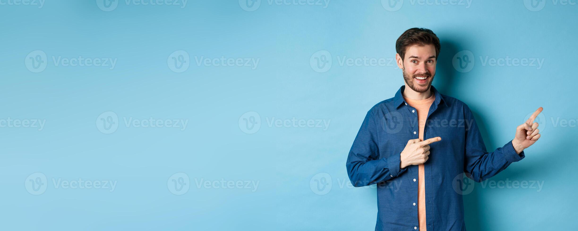 Cheerful caucasian man with beard pointing fingers aside at empty space and smiling at camera, showing advertisement, blue background photo