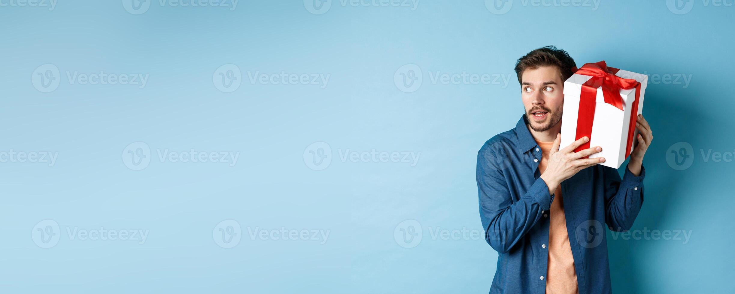 Valentines day. Curious boyfriend holding gift box near ear, trying to guess whats inside surprise present, standing over blue background photo