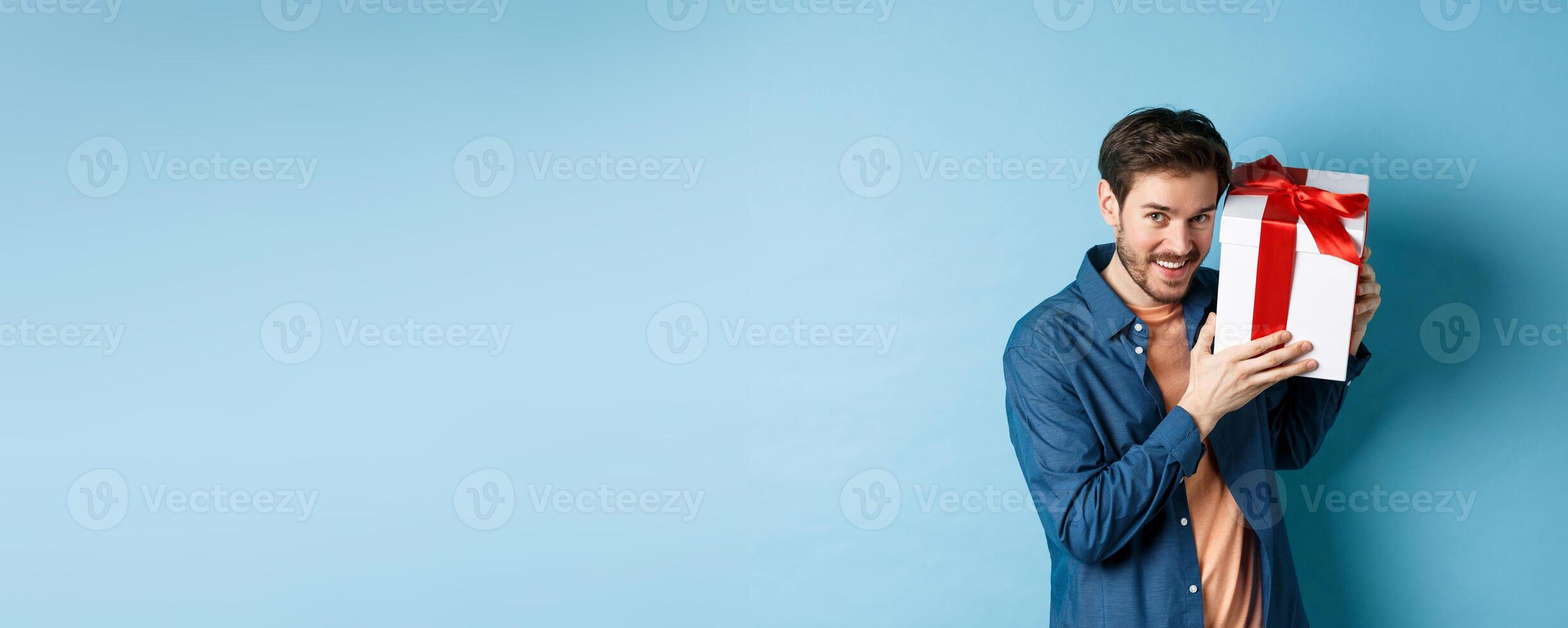 Valentines day. Romantic boyfriend showing gift box for lover, celebrating anniversary, standing over blue background photo