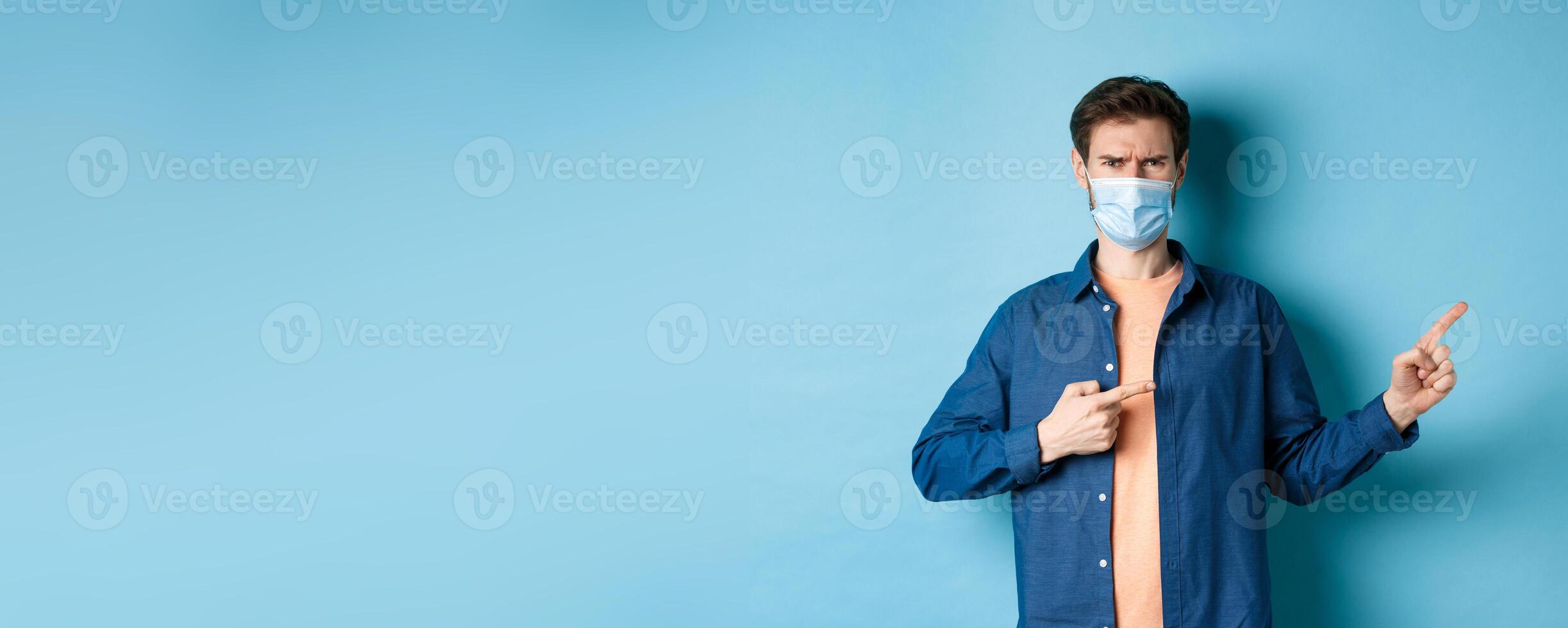 Covid-19 and pandemic concept. Angry man in medical mask frowning, pointig fingers at upper right corner empty space, complaining, standing on blue background photo
