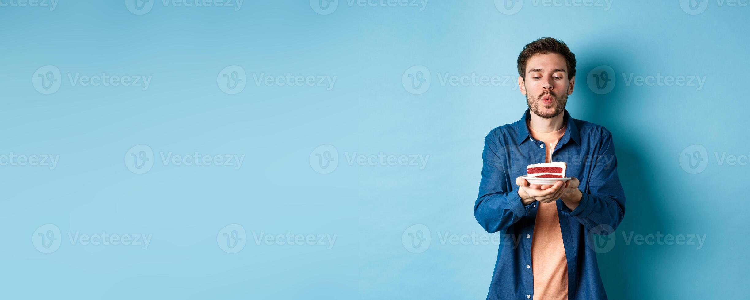 Celebration and holiday concept. Handsome young man blowing candle on birthday cake and making wish, standing on blue background photo