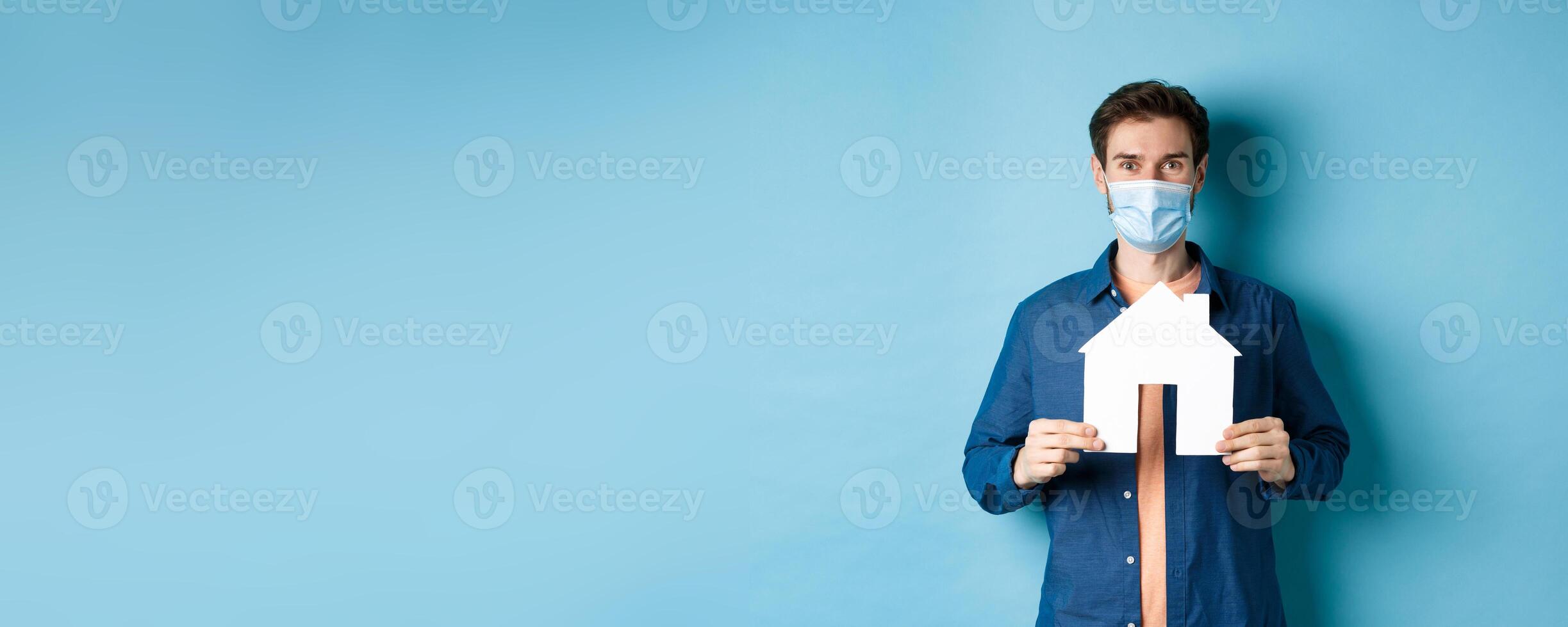 Real estate and quarantine concept. Happy young man in medical mask showing house cutout, buying property, standing on blue background photo