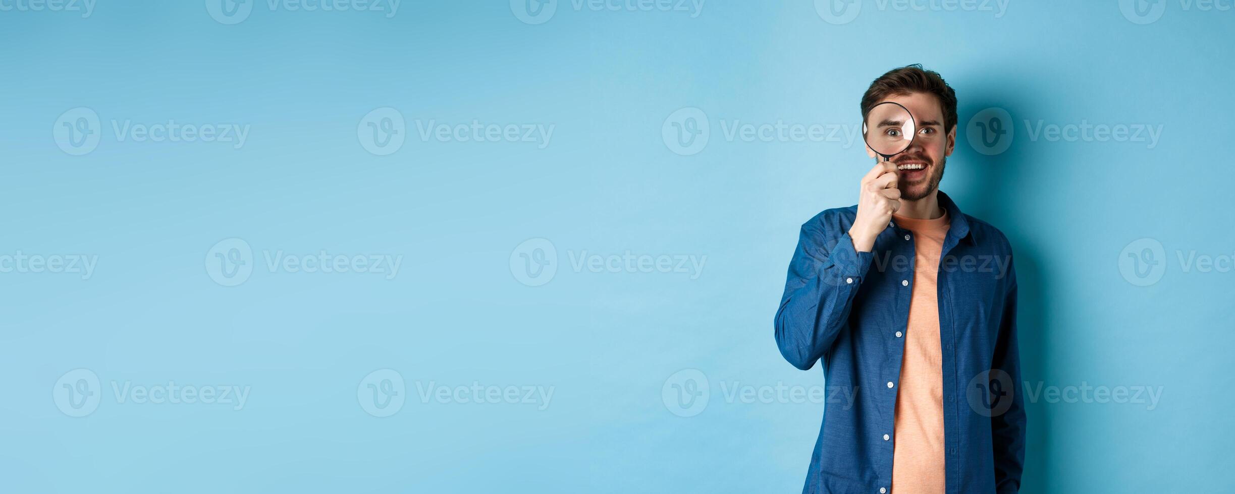 Cheerful young man found something good, smiling and looking through magnifying glass, standing on blue background photo