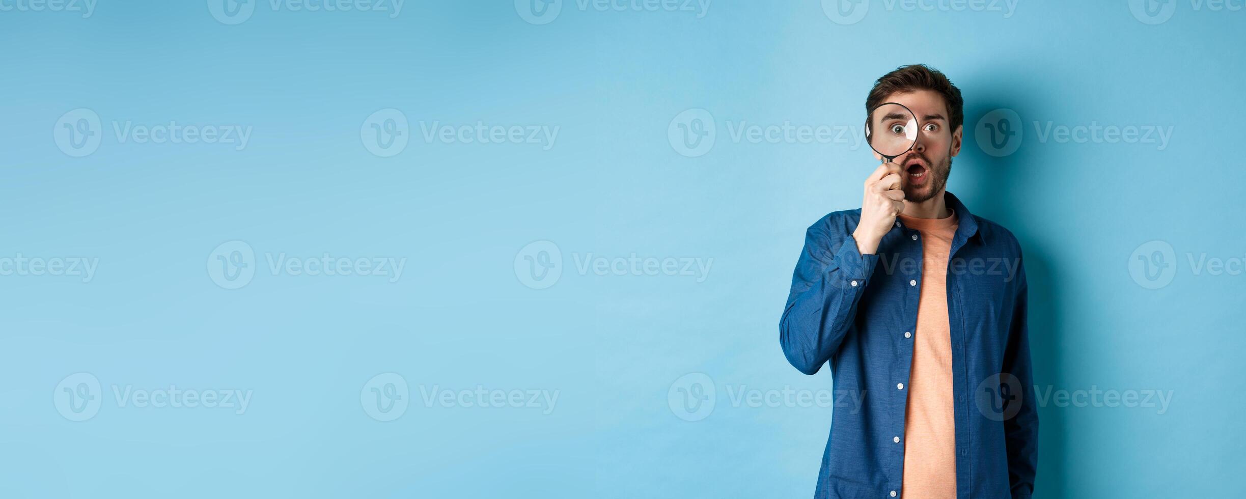 Impressed man look in awe through magnifying glass, drop jaw and stare at camera, standing on blue background photo