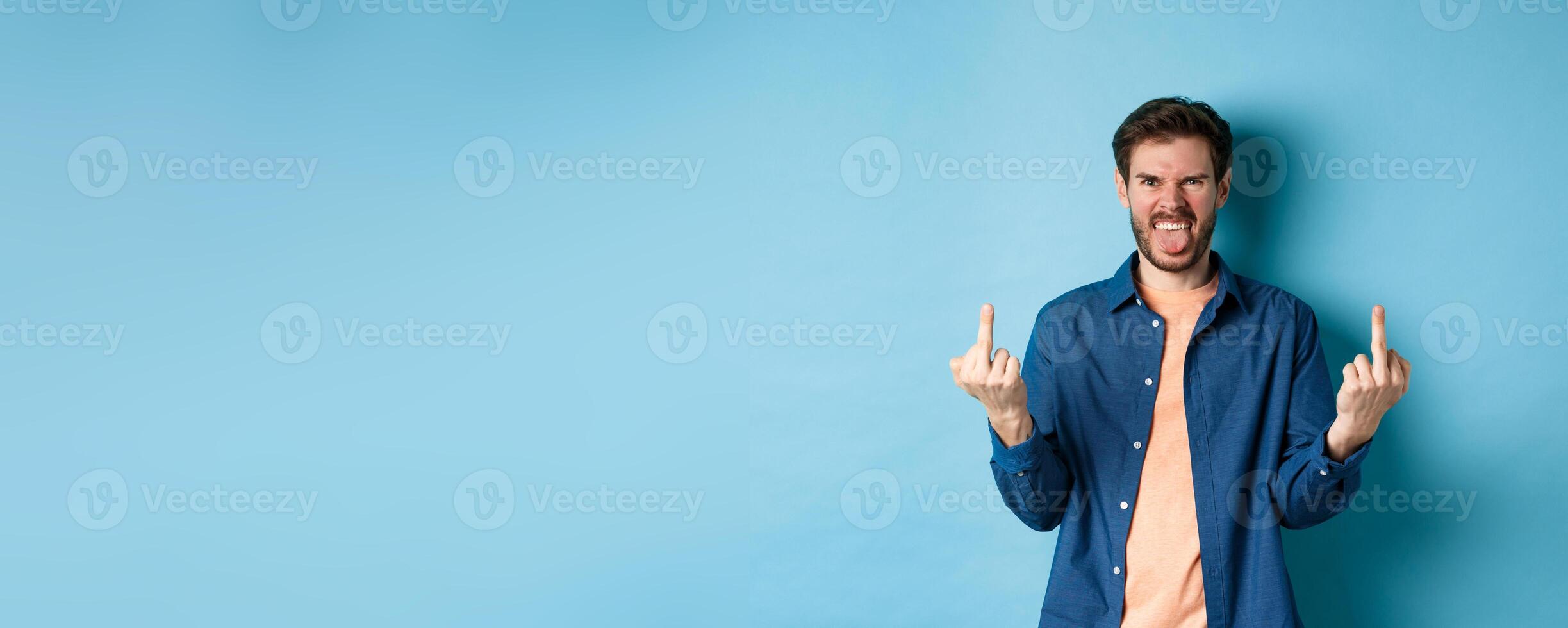 Ignorant and rude guy showing middle finger and smiling, say fuck you, standing on blue background photo