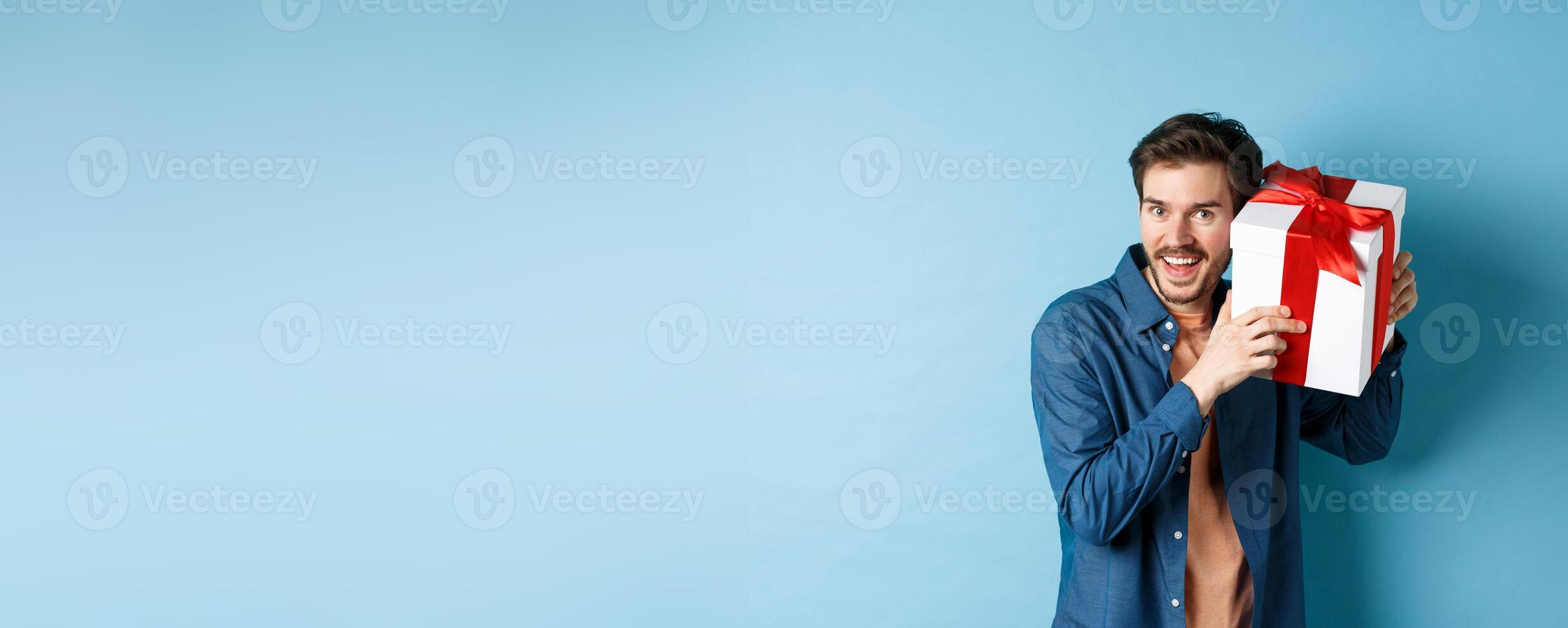Valentines day. Happy young man got present on special holiday, trying guess what inside gift box and smiling, standing over blue background photo
