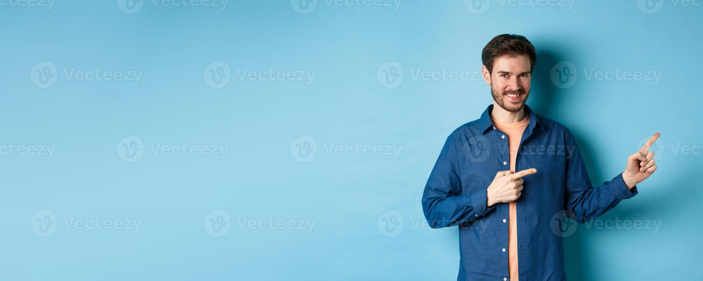 Smiling confident man showing logo, pointing fingers right at empty space and looking at camera, standing on blue background photo