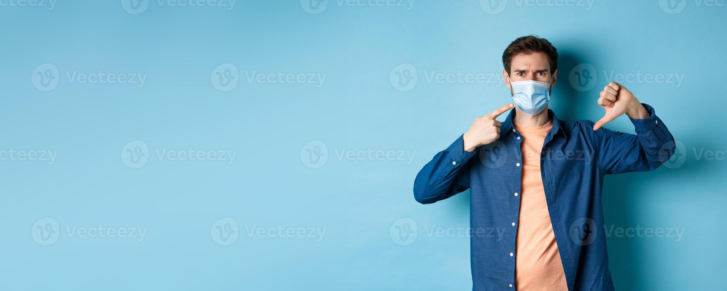 Coronavirus and pandemic concept. Disappointed young man pointing at medical mask and showing thumb down, disapprove and dislike quarantine rules, blue background photo