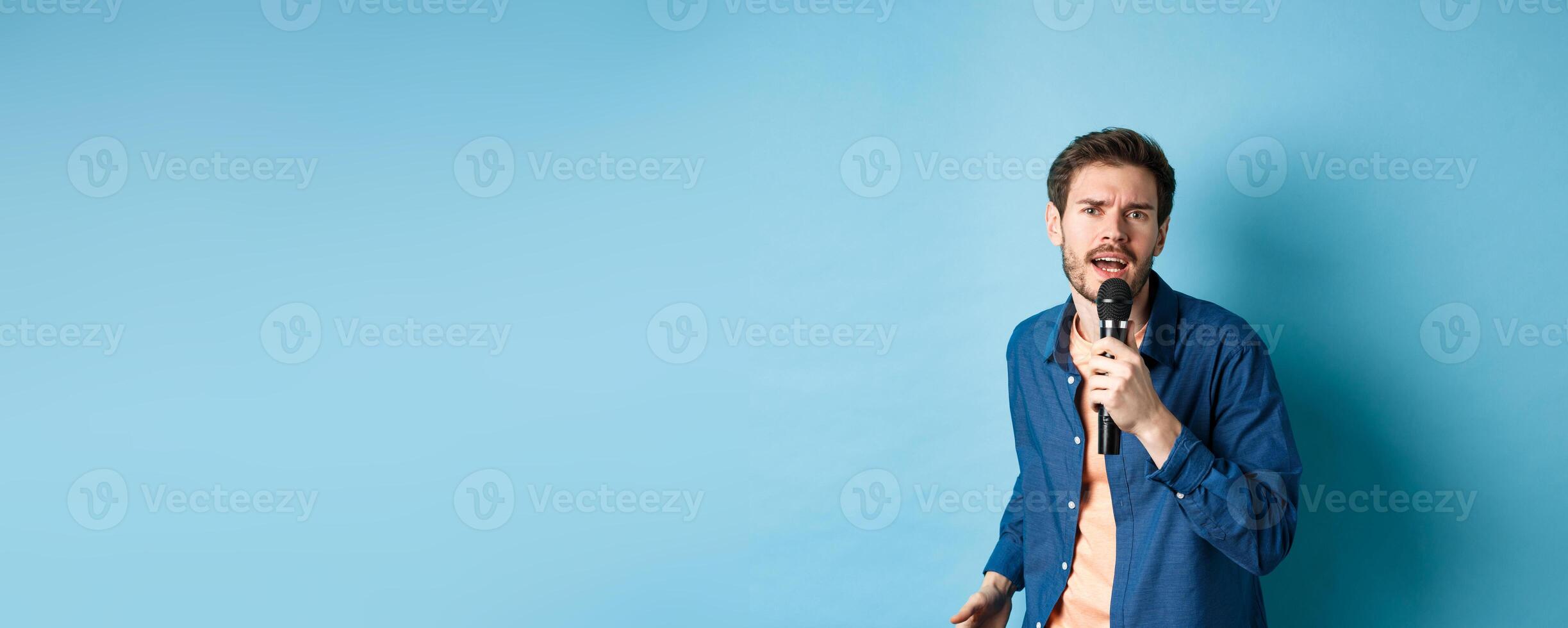 Passionate singer looking at camera, singing in microphone, playing karaoke on blue background photo