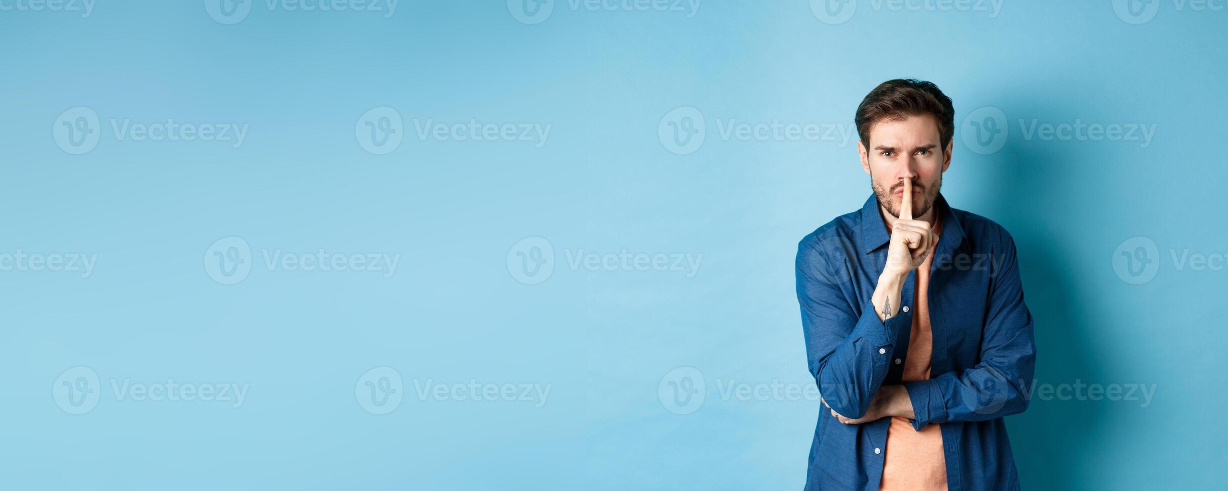 Angry man shushing, press finger to lips and frowning, tell to be quiet, show taboo gesture, standing on blue background photo