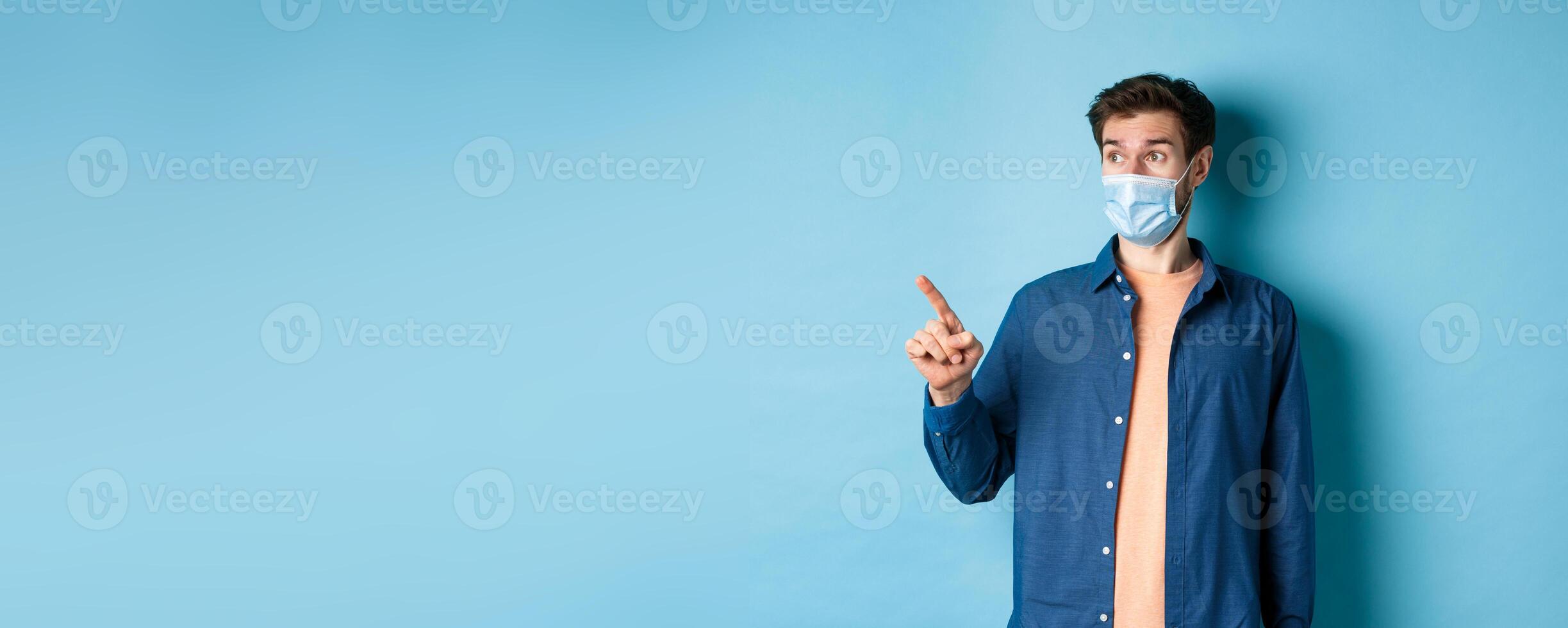 Covid-19 and healthcare concept. Surprised guy in face mask pointing and looking left at empty space, standing on blue background photo