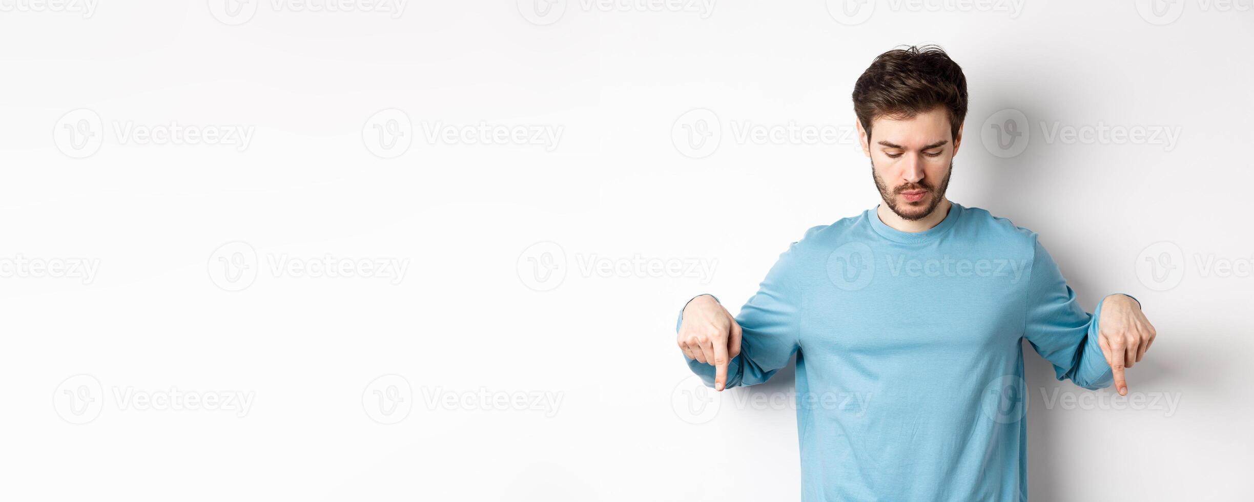 Handsome caucasian man with beard, wearing blue shirt, looking and pointing down at interesting logo banner, standing on white background photo