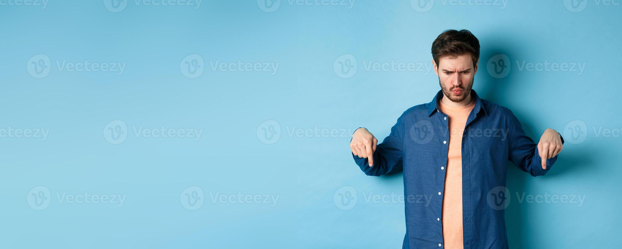 Confused and shocked guy looking and pointing down at something strange, standing on blue background photo