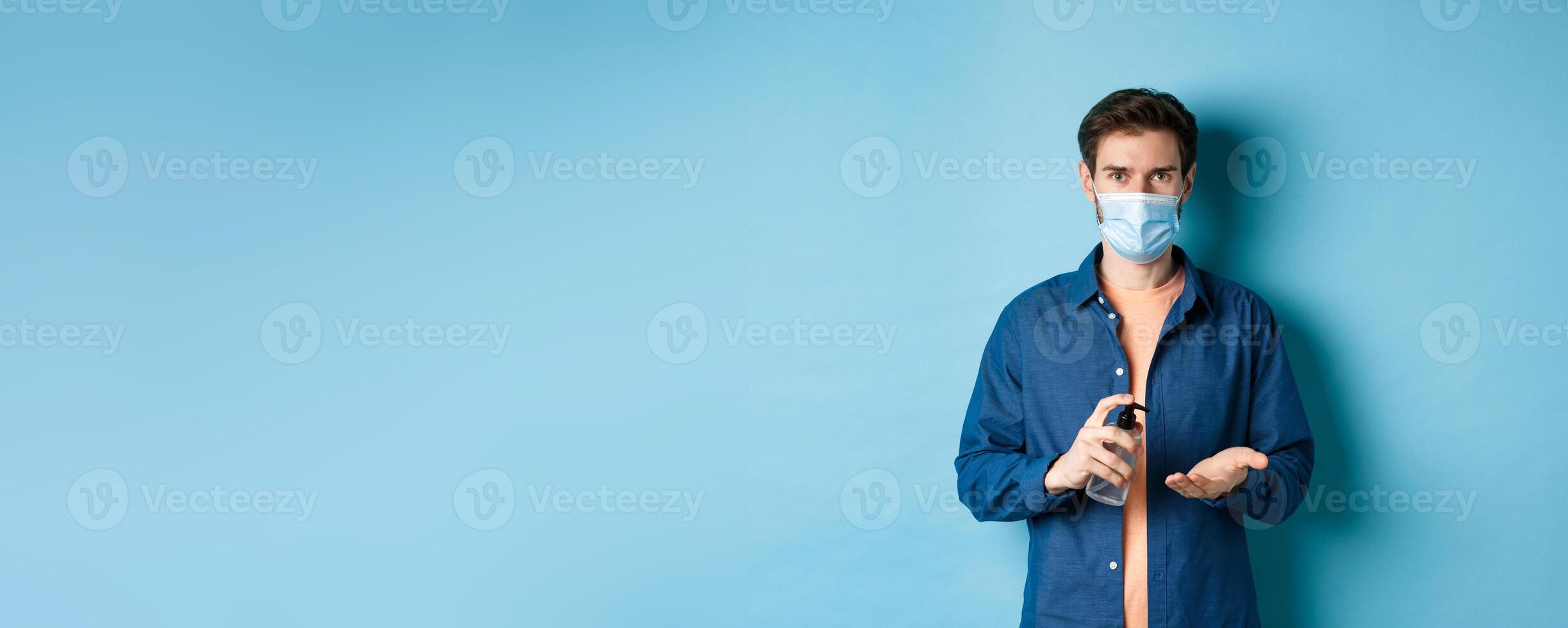 Covid-19, health and pandemic concept. Young guy in medical mask cleaning hands with sanitizer, apply antiseptic on palm, standing on blue background photo