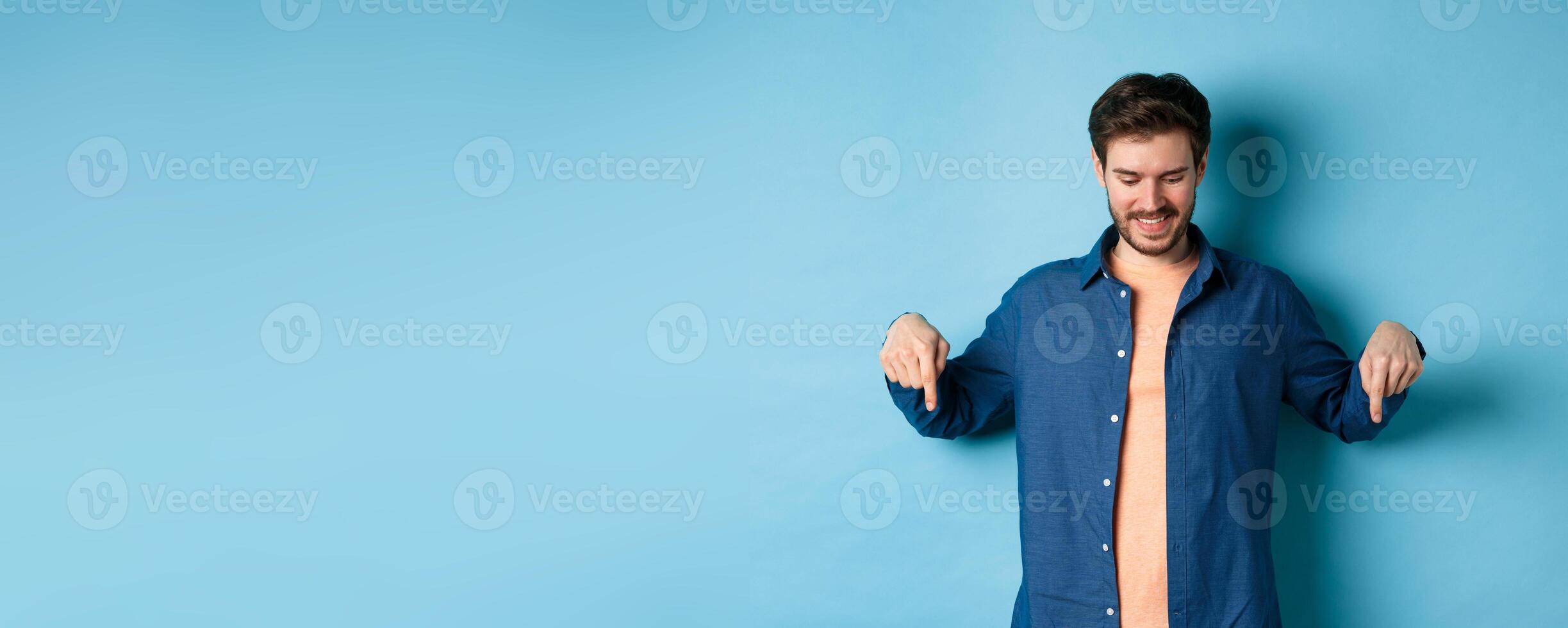Smiling handsome man with beard, looking and pointing down at banner, checking out special offer, standing on blue background photo