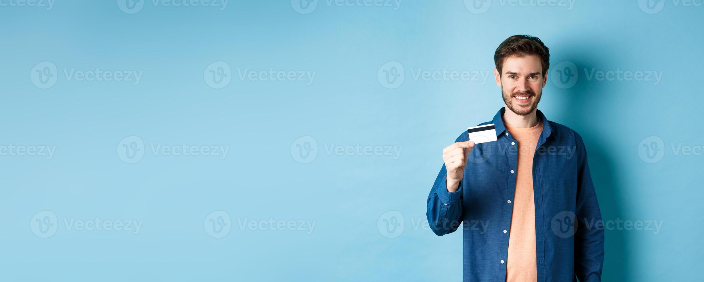 Handsome smiling guy showing plastic credit card and looking satisfied, standing on blue background photo