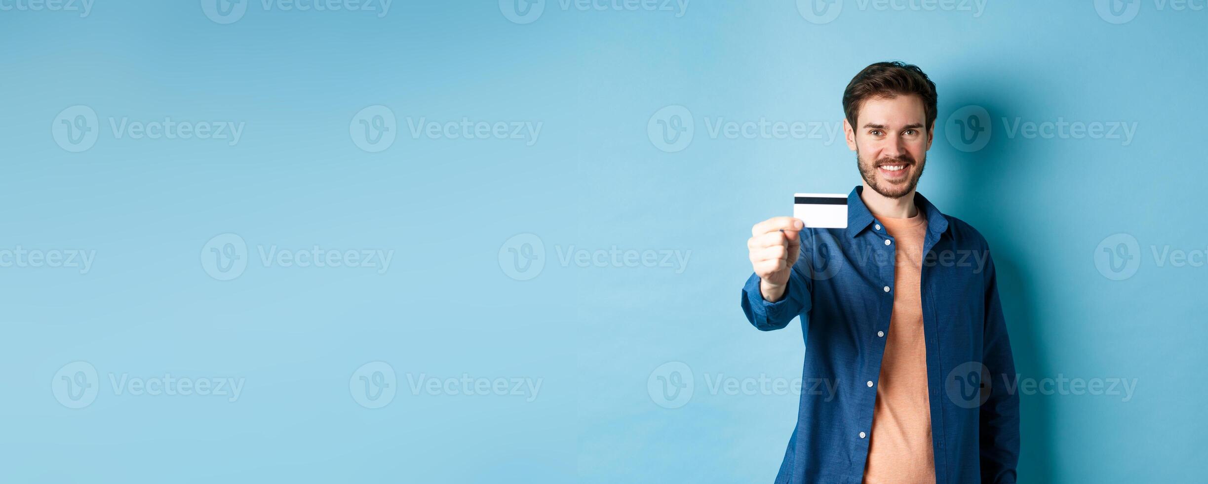 Cheerful caucasian man showing plastic credit card and smiling, recommending bank, standing on blue background photo